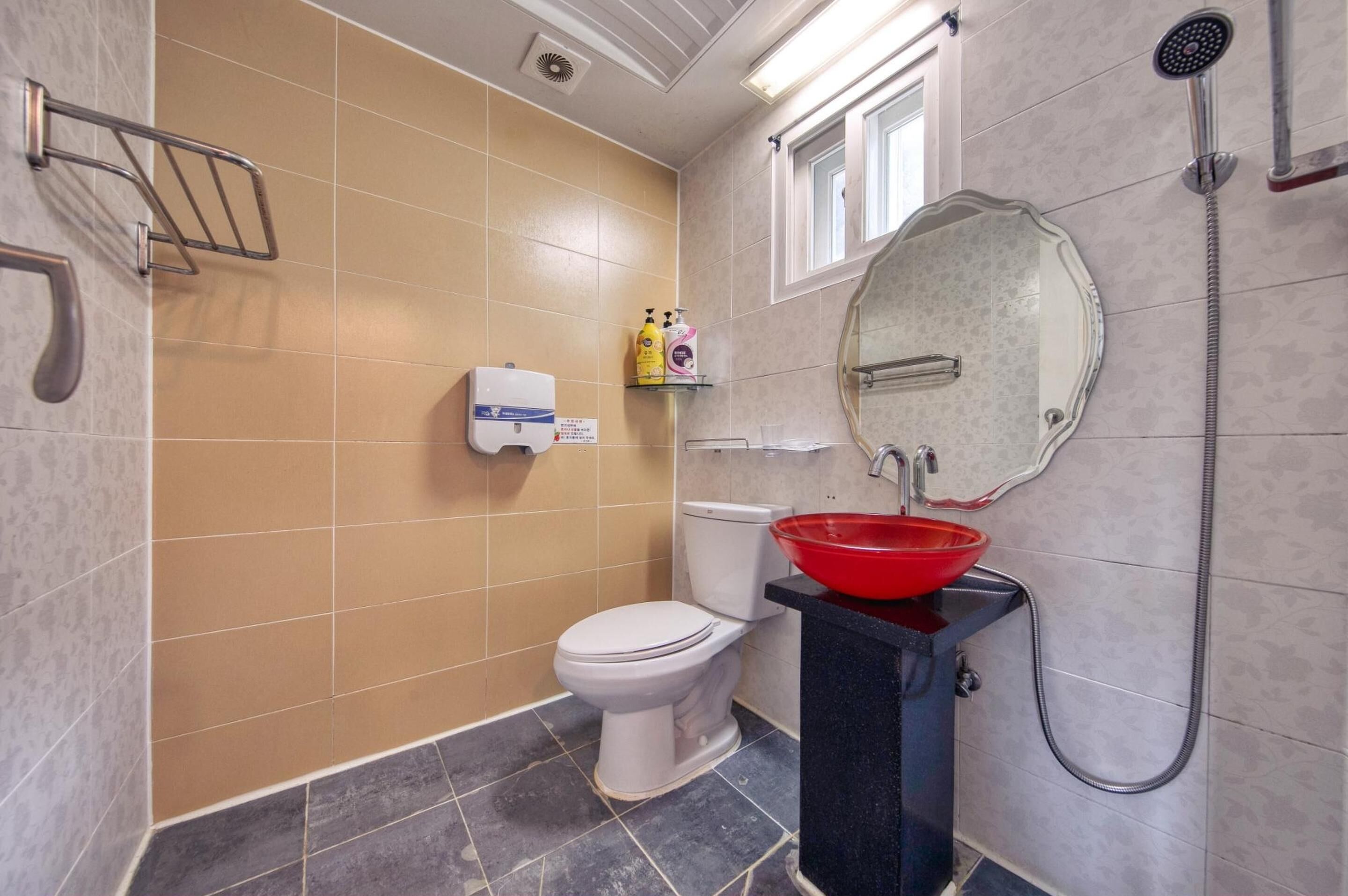 Une salle de bain comprenant des toilettes, un lavabo avec un récipient rouge, une douchette à main et un miroir avec un cadre décoratif.
