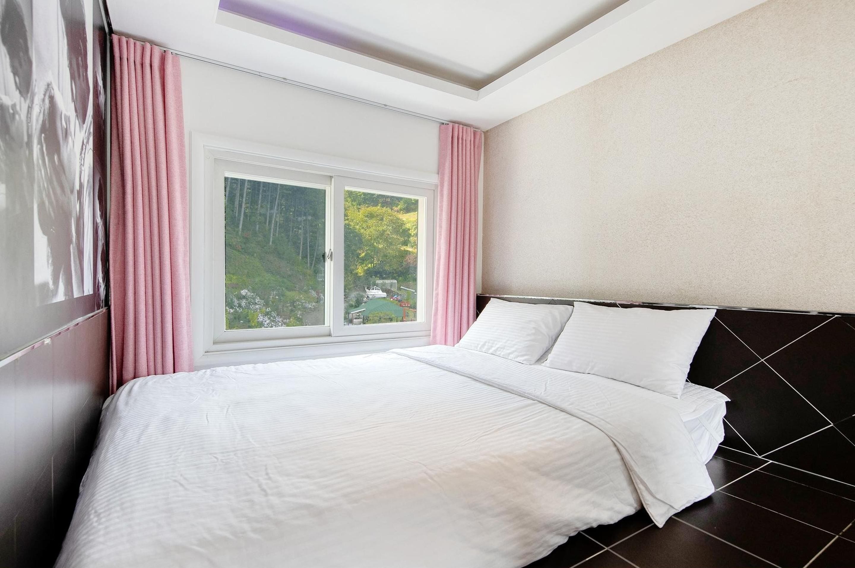 A bedroom with a large bed, a window with curtains, and a view of greenery outside.