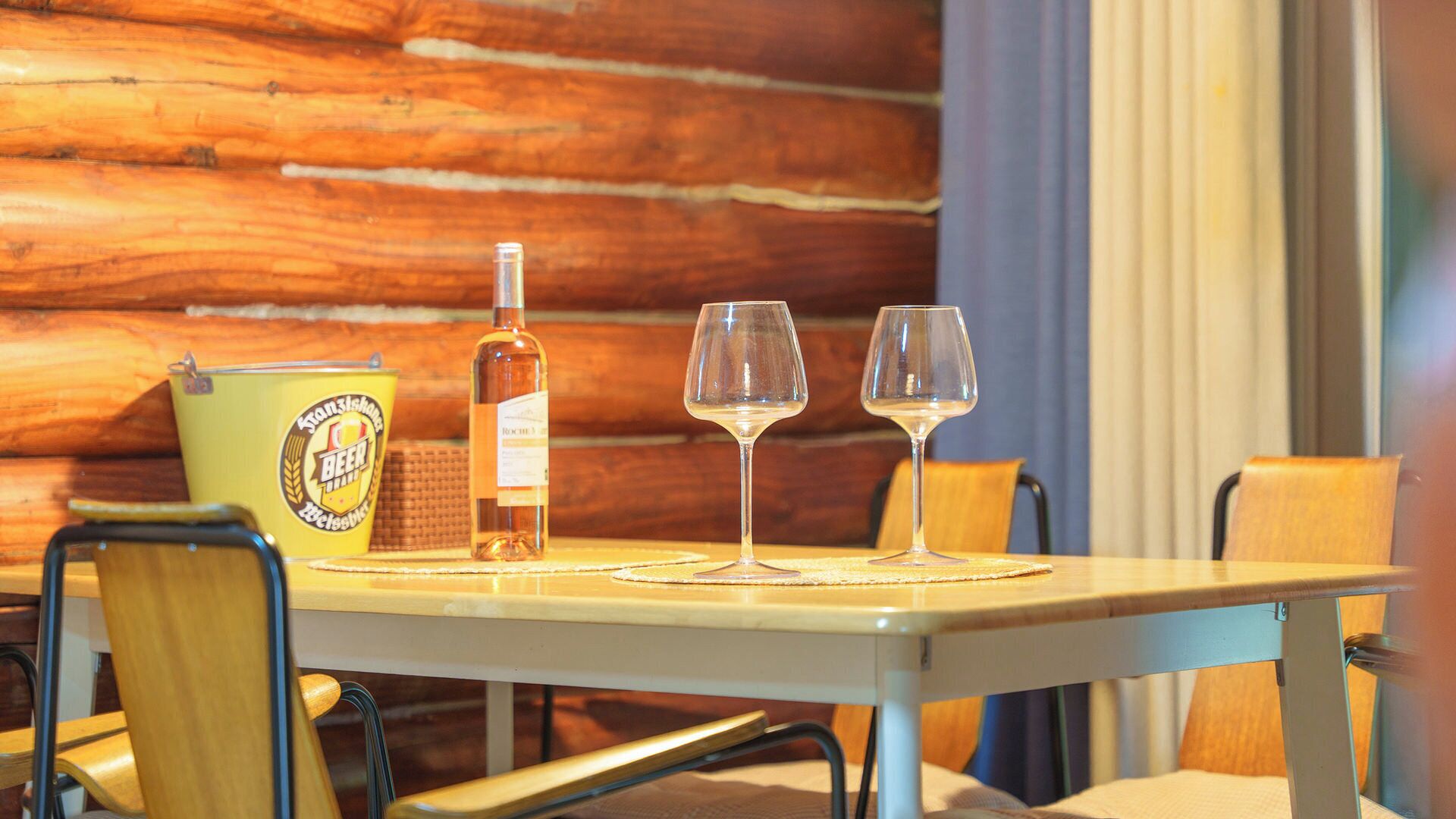Un interior de madera con una mesa preparada para dos personas, incluyendo una botella y dos vasos.