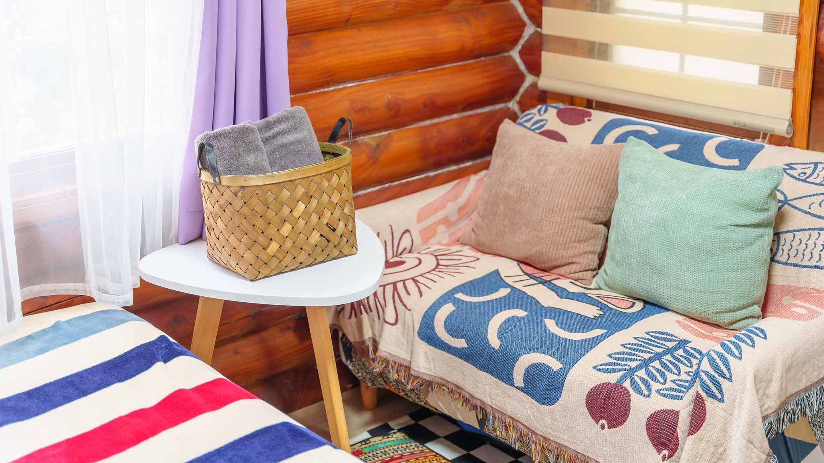 A cozy cabin interior with a woven basket on a small table, a sofa with patterned cushions, and a striped blanket on the floor.