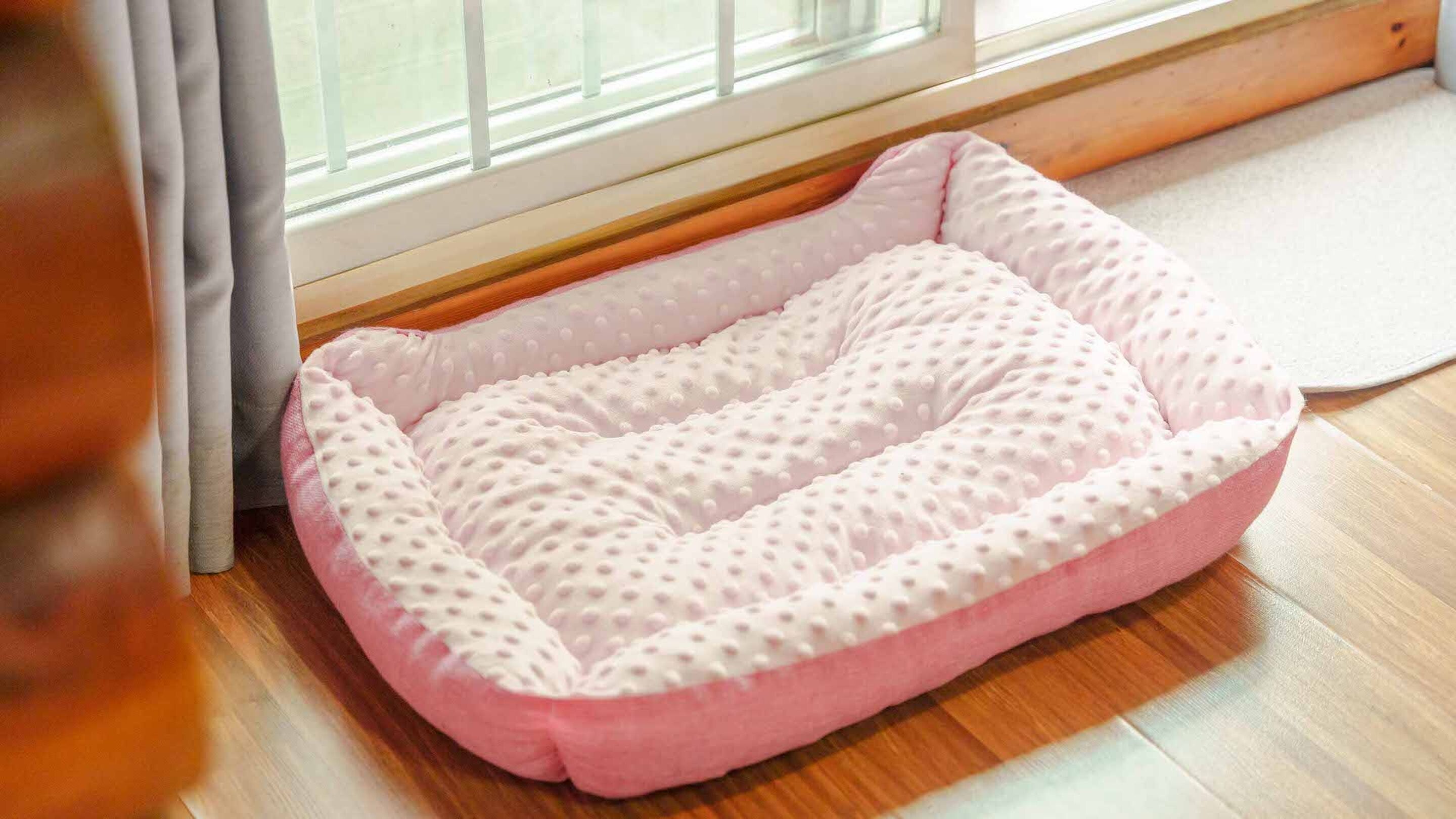 Ein pinkfarbenes Haustierbett mit strukturierter Oberfläche auf einem Holzboden in der Nähe eines Fensters mit weißem Vorhang.