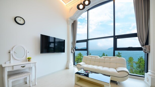 Un salon moderne comprenant un canapé, un téléviseur, une coiffeuse avec un miroir et une grande fenêtre donnant sur la mer.