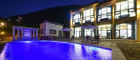 A swimming pool with a blue-lit edge, surrounded by lounge chairs and umbrellas, with a building featuring large windows and outdoor seating in the background.