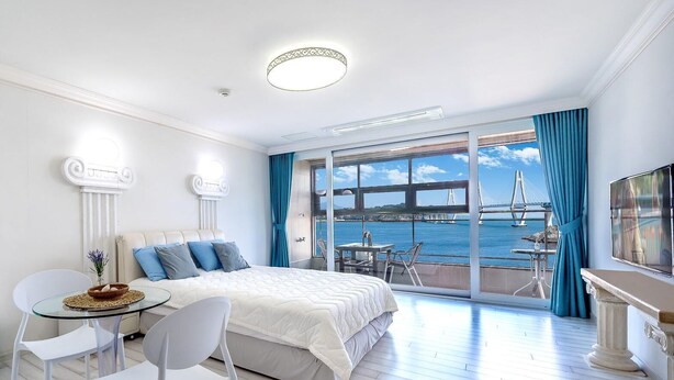 A modern hotel room with a large bed, a balcony with a view of a bridge and water, and a dining area with a table and chairs. - Geoje Healing Pension (Geoje)