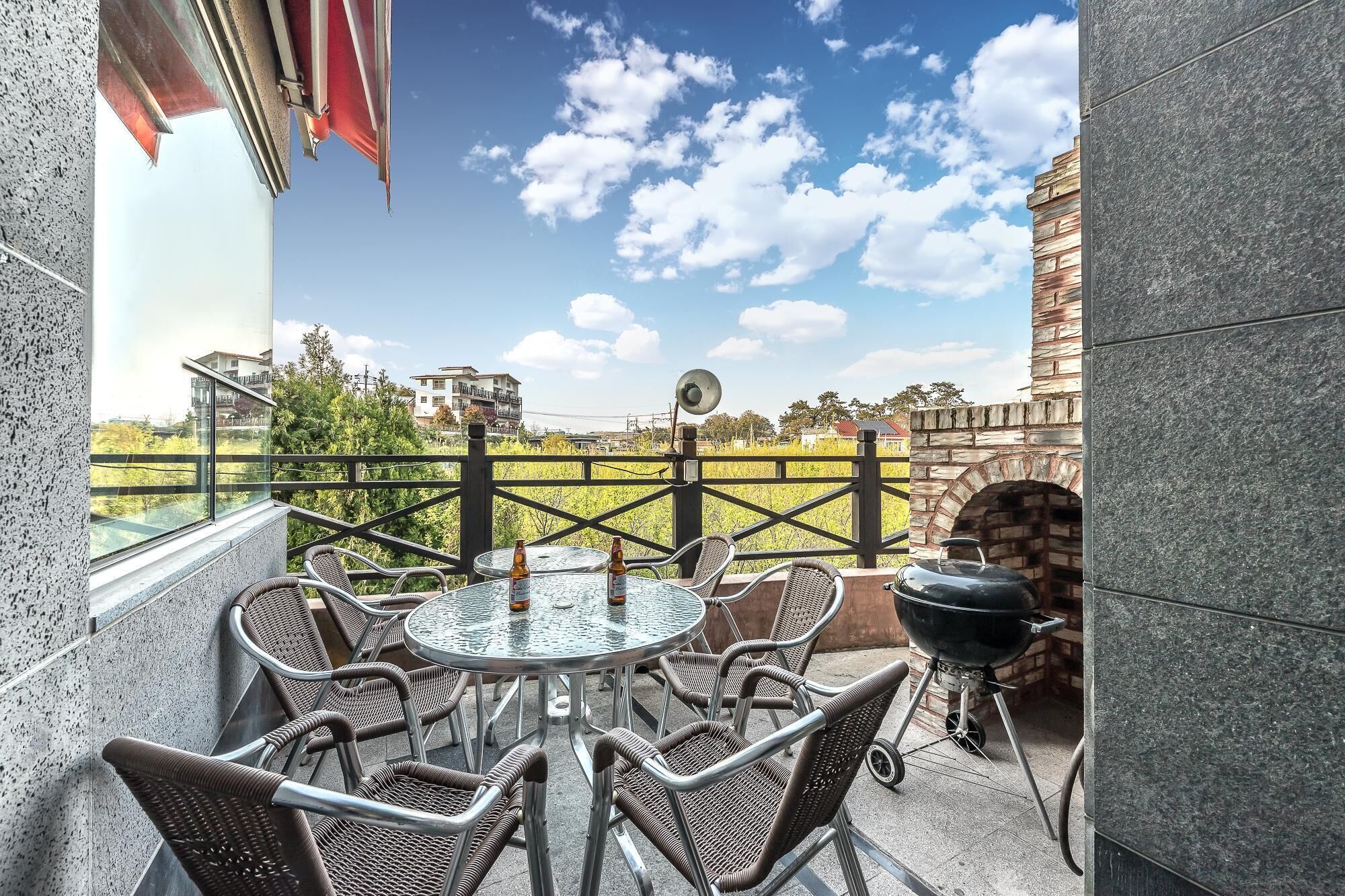 A balcony with wicker chairs and a round glass table, a barbecue grill, and a view of a brick building and green fields.