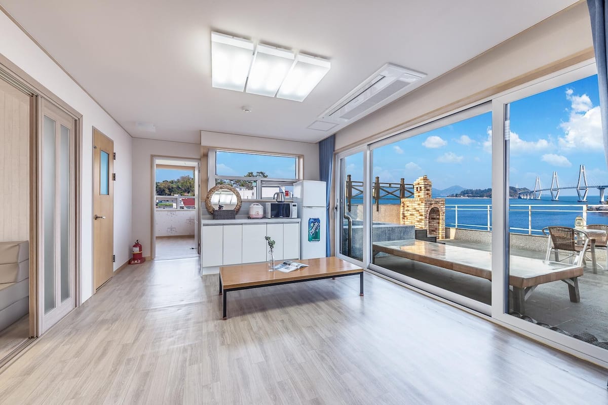 A modern room with a wooden floor, a kitchen area with a refrigerator, and a large sliding glass door leading to a balcony with a view of a bridge and water.
