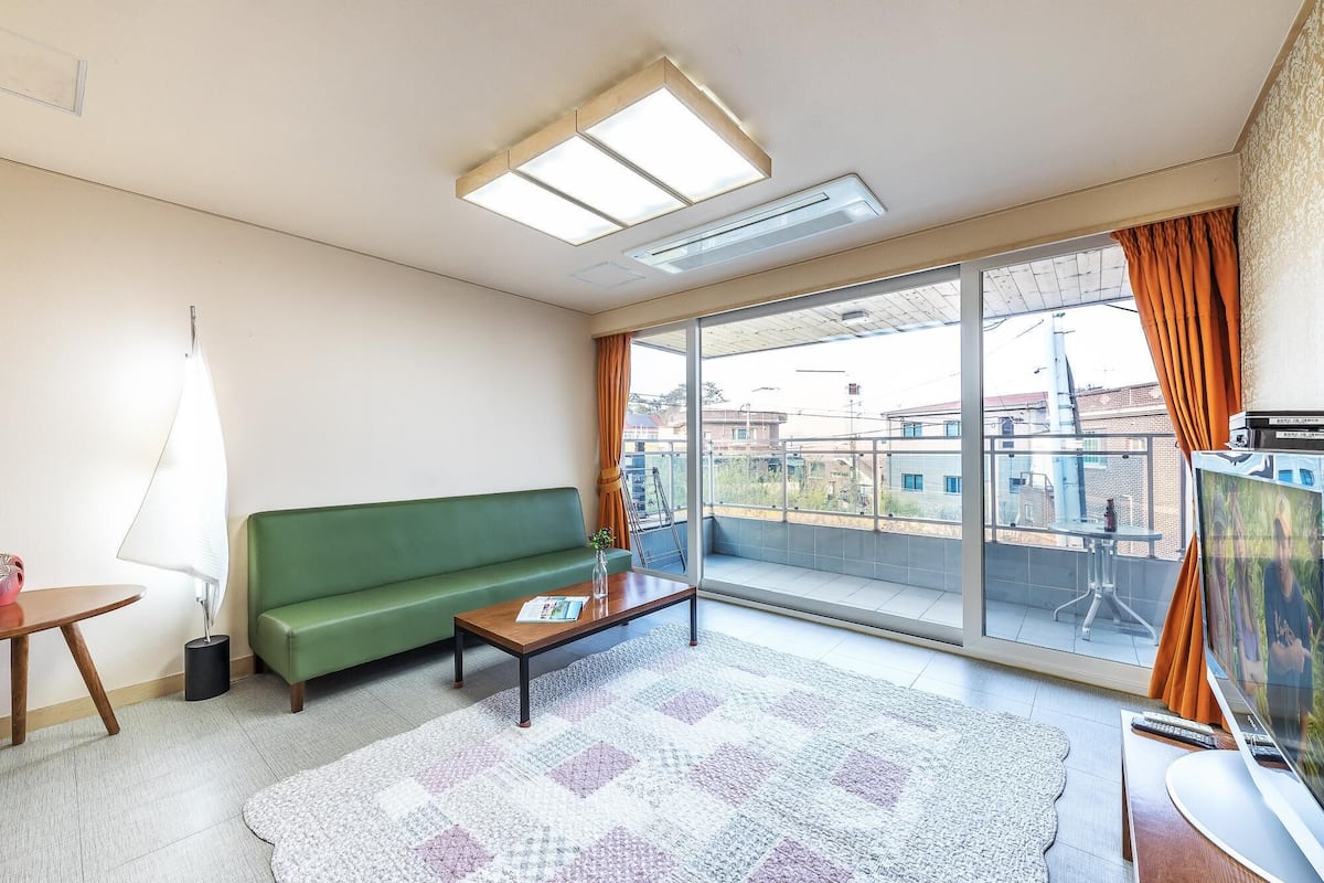 A living room with a green sofa, a wooden coffee table, a patterned rug, and a large sliding glass door leading to a balcony.