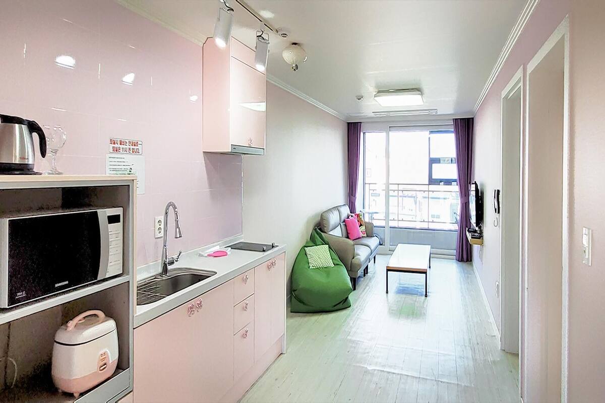 A compact kitchen with a microwave, sink, and a small countertop. There is a green bean bag chair and a sofa with cushions in the living area. The room has a balcony with a view.