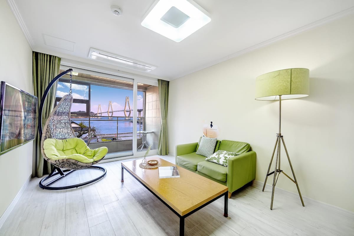 A modern hotel room with a green sofa, a hanging chair, a coffee table, and a floor lamp. A large window offers a view of a bridge and water.
