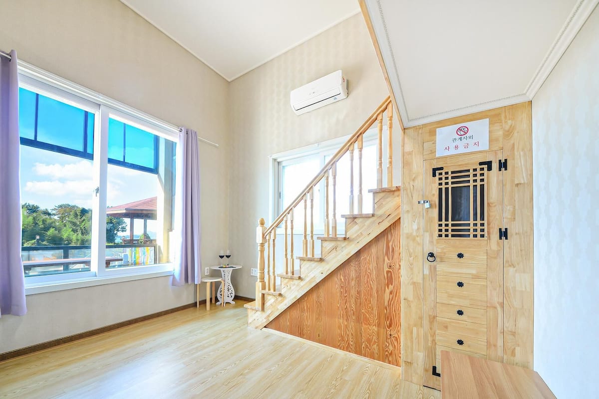 Ein Zimmer mit einer Holztreppe, großen Fenstern mit Vorhängen, einem kleinen Tisch mit Stuhl und einer Tür mit einem Schild.