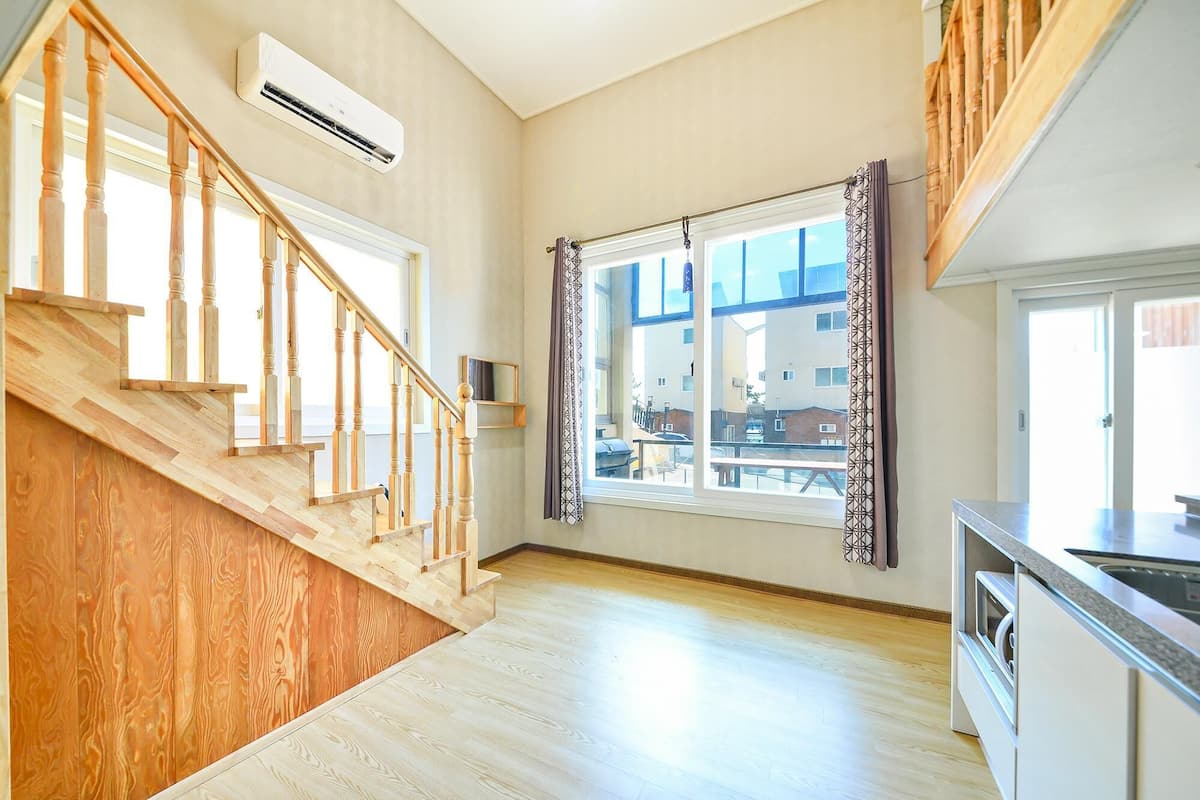 Ein geräumiges Zimmer mit einer Holztreppe, einem großen Fenster mit Vorhängen und einer Küchenzeile.