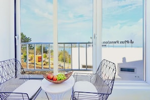 A balcony with a table and two chairs, a bowl of fruit, and a view of the sea and buildings. - Yeosu Jplus A Pension (Yeosu)
