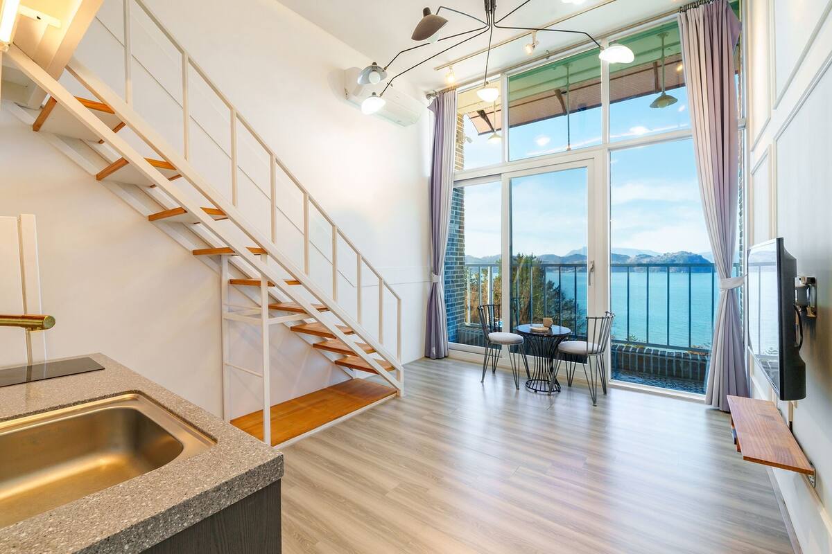Eine moderne Wohnung mit Küche, einer Holztreppe, einem Essbereich mit Meerblick und einem Balkon mit Fernseher.