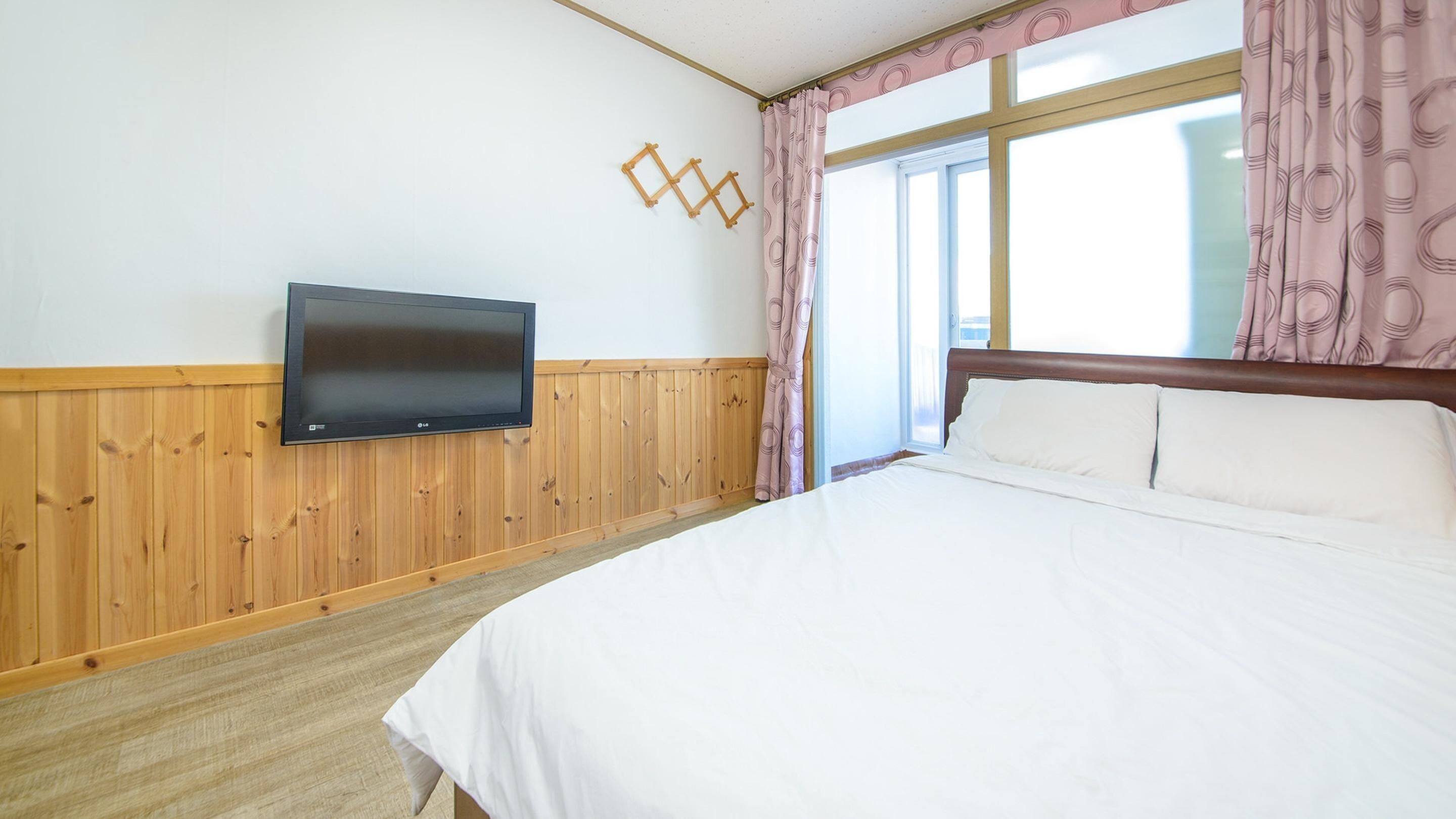 A bedroom with a bed, a flat-screen TV mounted on a wooden wall, and a window with curtains.