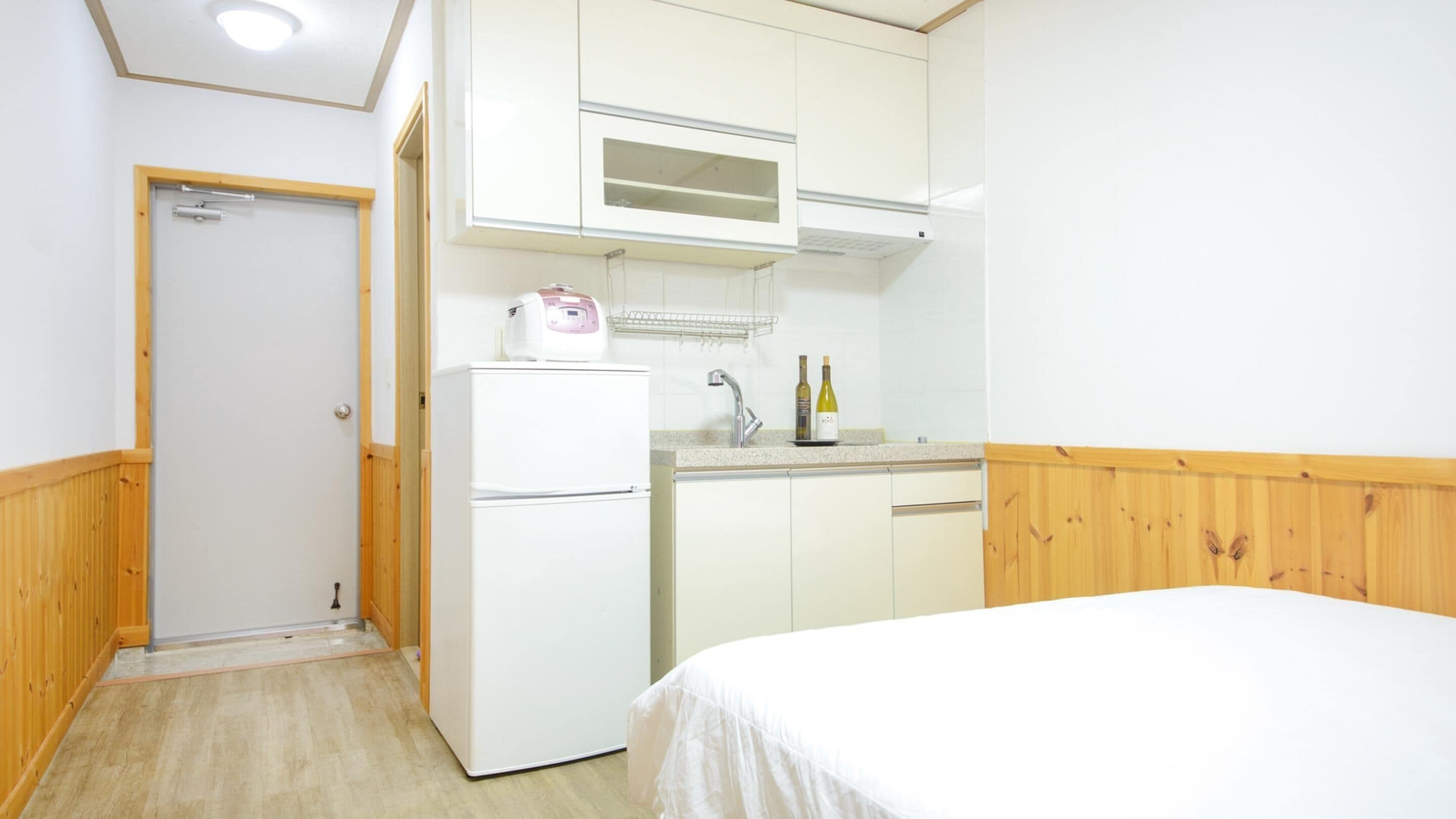 Ein kleines Zimmer mit einem Bett, einem weißen Kühlschrank, einer kleinen Küchenzeile mit einer Mikrowelle und einer Holztür.