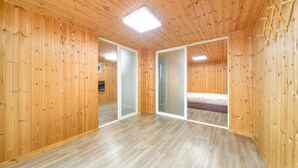 A room with wooden walls and a wooden floor, featuring a bed, a small seating area, and a sliding glass door leading to another room. - Samcheok sky Pension (Samcheok)