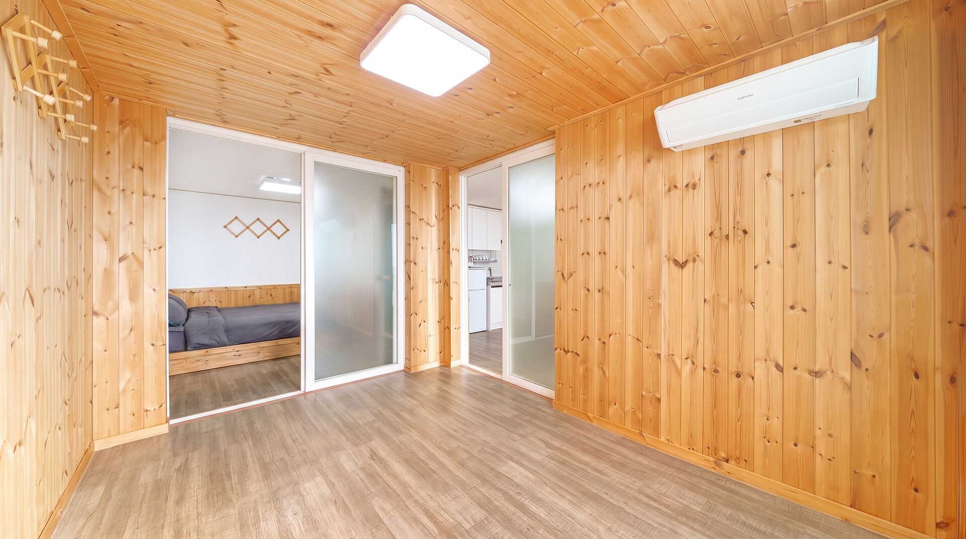 A room with wooden walls and a ceiling, a bed, a sliding door, and a kitchen area visible through the door.