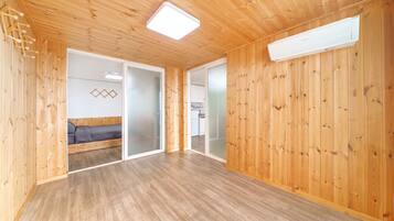 A room with wooden walls and a ceiling, a bed, a sliding door, and a kitchen area visible through the door.