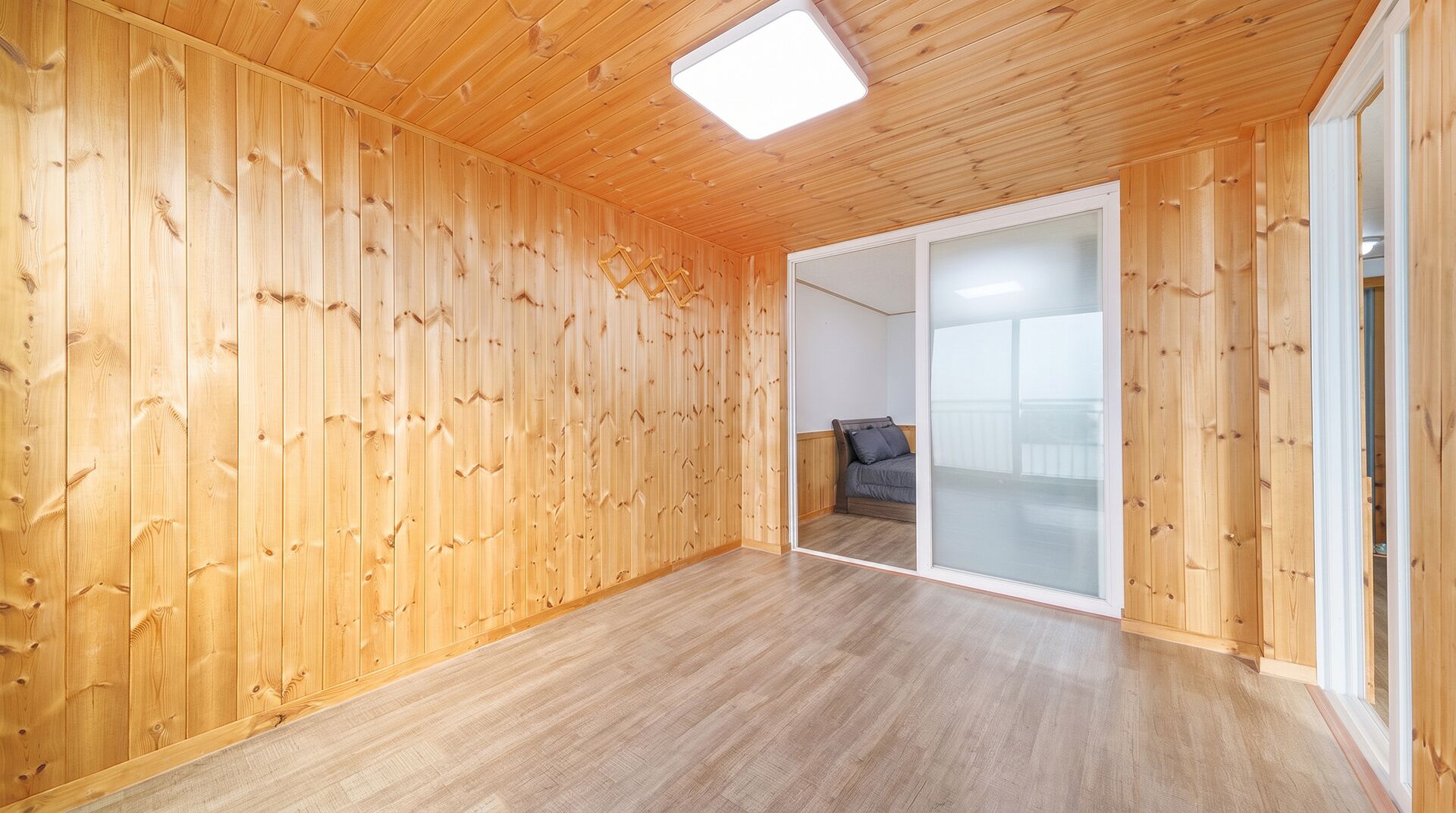 A room with wooden walls and a wooden floor, a sliding glass door leading to another room, and a ceiling light.