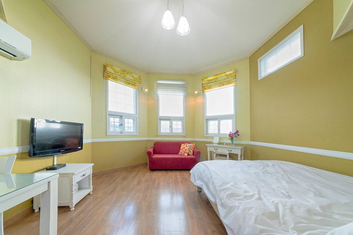 A hotel room with a bed, a TV, a red sofa, a small table, and two windows with curtains.