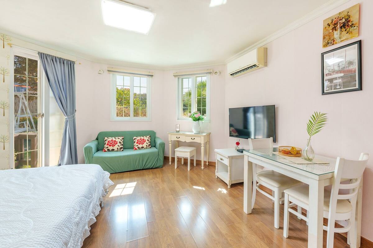 A bedroom with a bed, a green sofa, a desk with a chair, a TV, and a window with curtains.