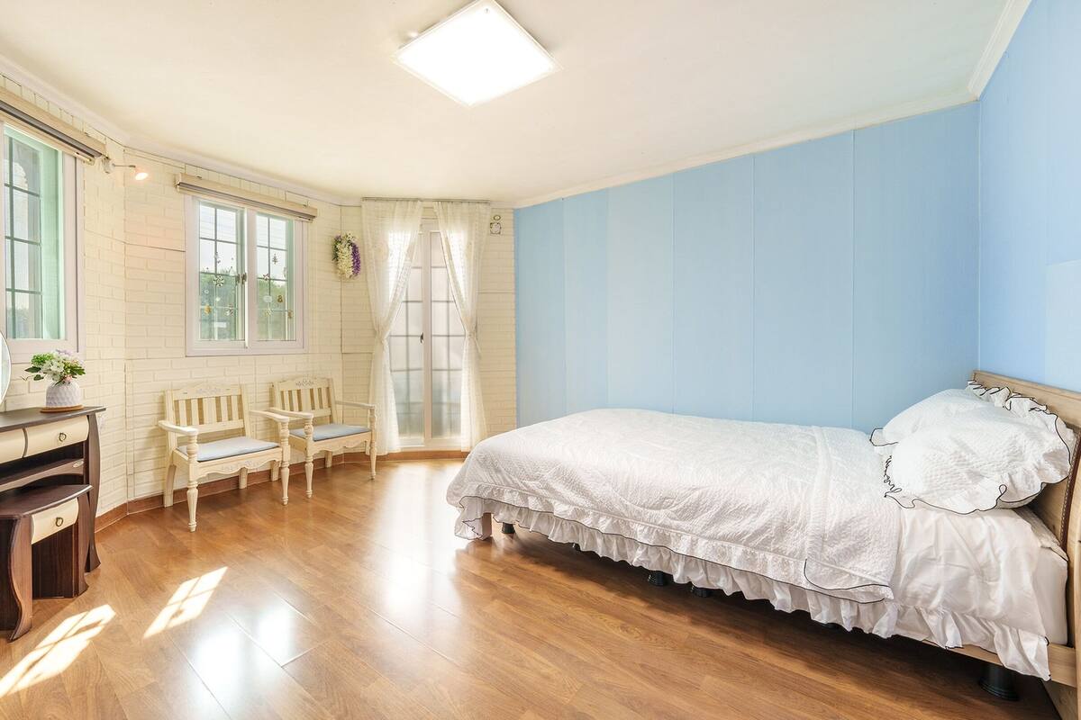 A bedroom with a wooden floor, a bed with white bedding, a dresser, two chairs, and a window with curtains.