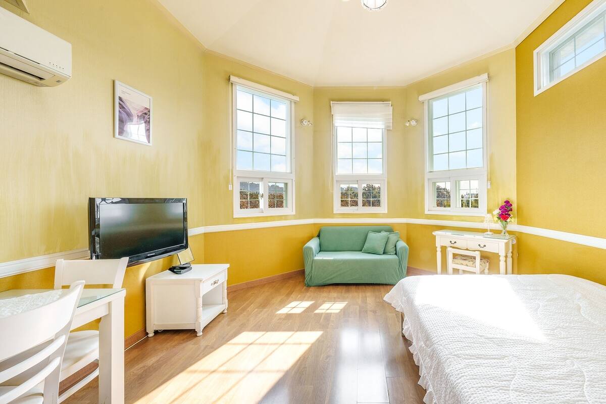 A bright yellow room with a bed, a green sofa, a TV, and a desk with a vase of flowers.