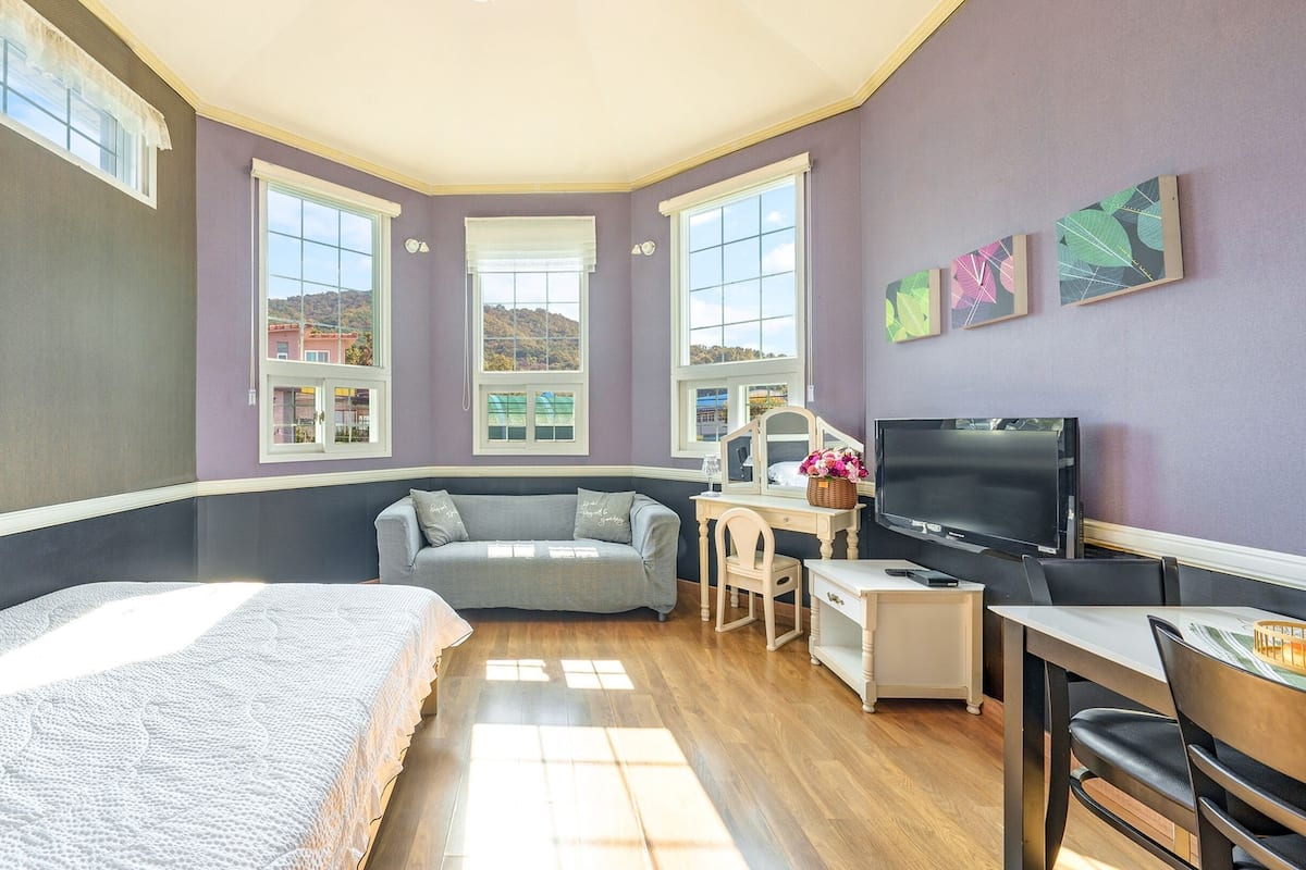 A room with a bed, a desk, a TV, and a couch. There are three windows providing natural light.