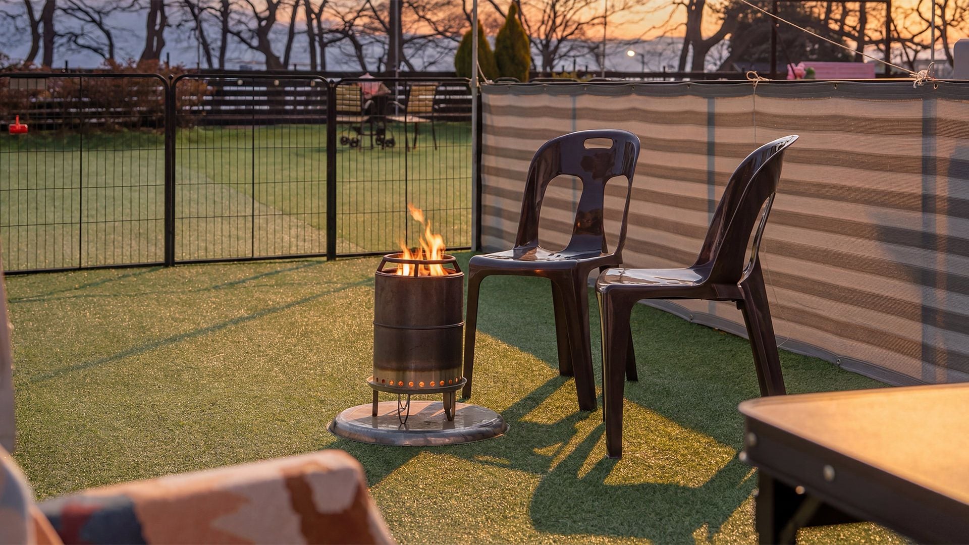 Un área de patio con césped artificial, un brasero metálico con llamas y dos sillas de plástico.