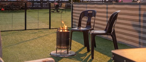 A patio area with artificial grass, a metal fire pit with flames, and two plastic chairs.