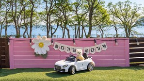 A white convertible car parked in front of a pink backdrop with a large white flower and Korean text.