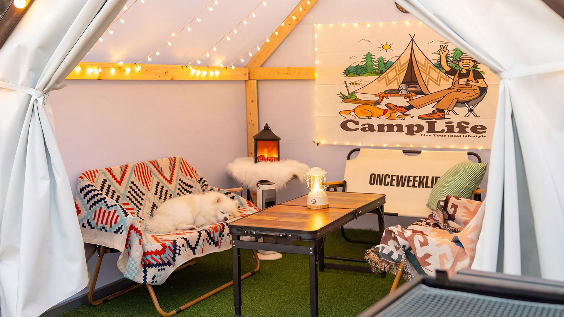 Un espace de camping intérieur accueillant, avec un coin nuit, une table avec une lanterne et une tente ornée d’une pancarte « CampLife ».