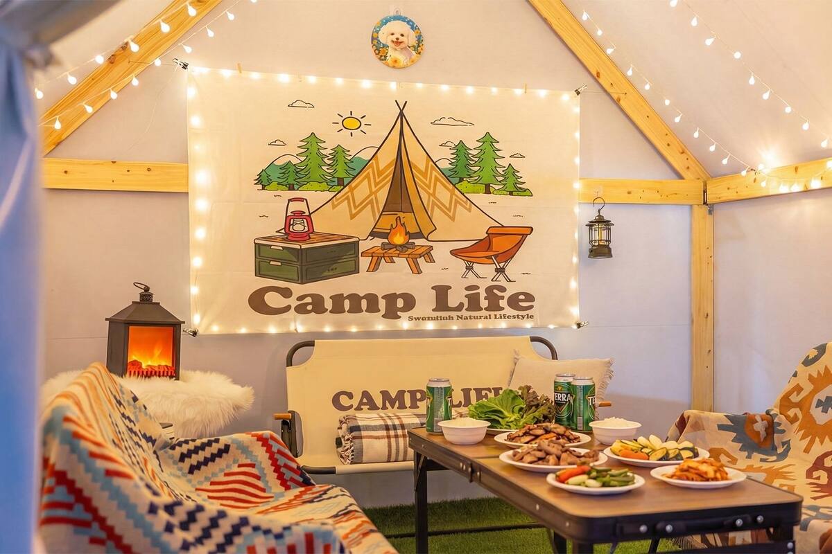 A cozy tent interior with a 'Camp Life' banner, a campfire, and a table with food.