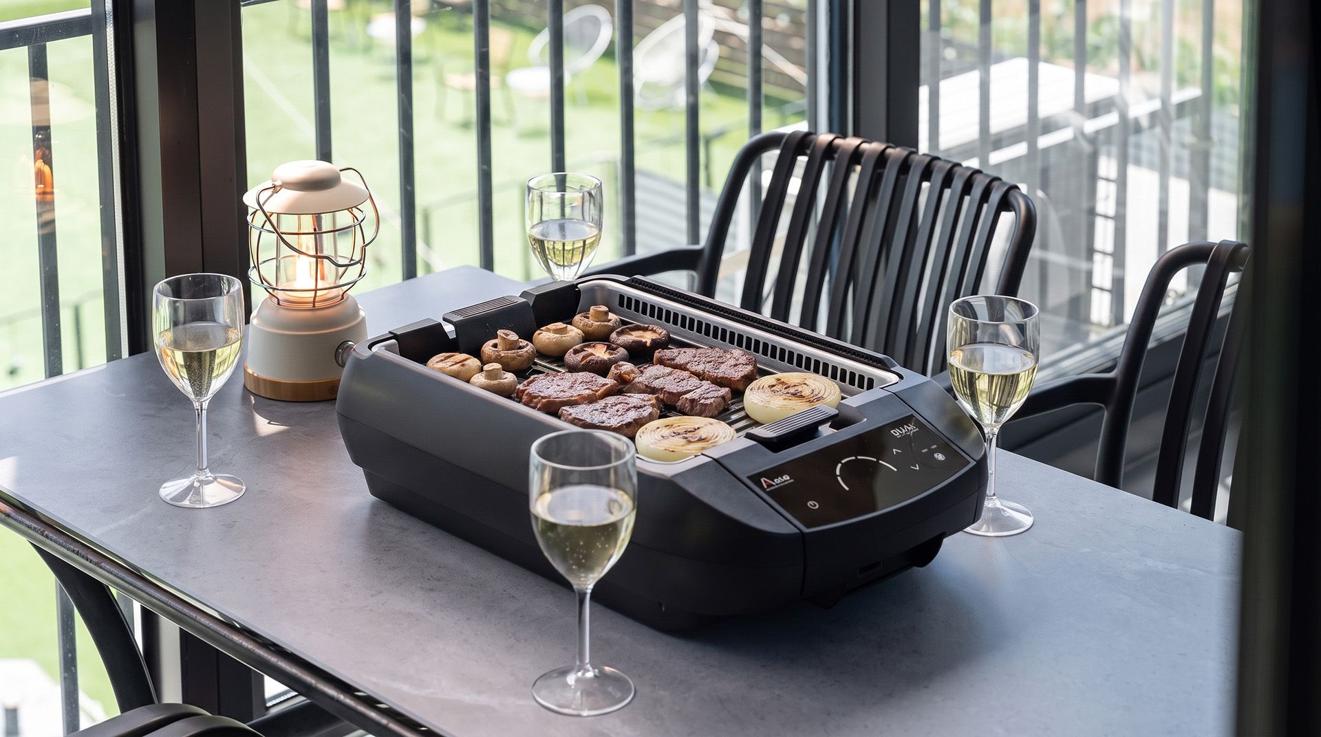 A table with a portable grill, grilled meat, mushrooms, and onions, accompanied by wine glasses.