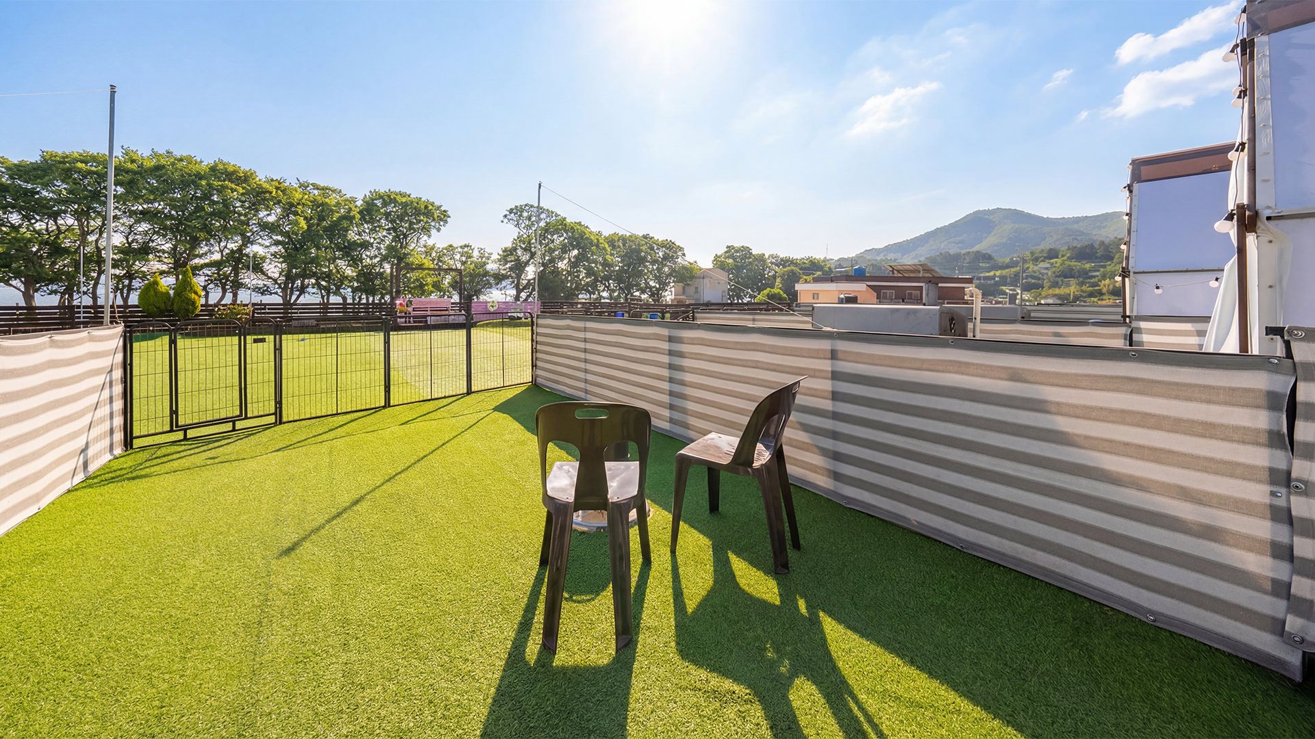 A rooftop terrace with artificial grass, two chairs, a metal railing, and a view of a mountain.