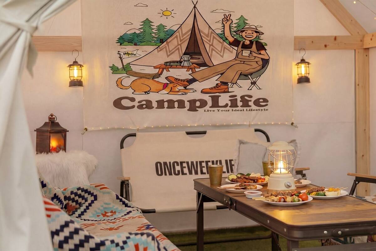 A cozy indoor camping setup with a tent, a person relaxing, and a table with food.