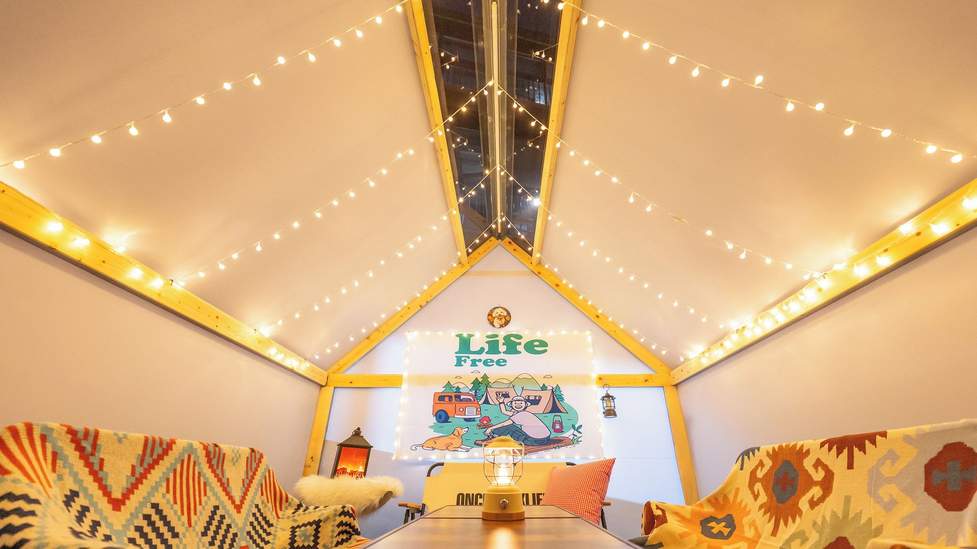 Una habitación acogedora con techo inclinado, luces colgantes, un mural con la inscripción 'Life Free', un farol y cojines estampados.