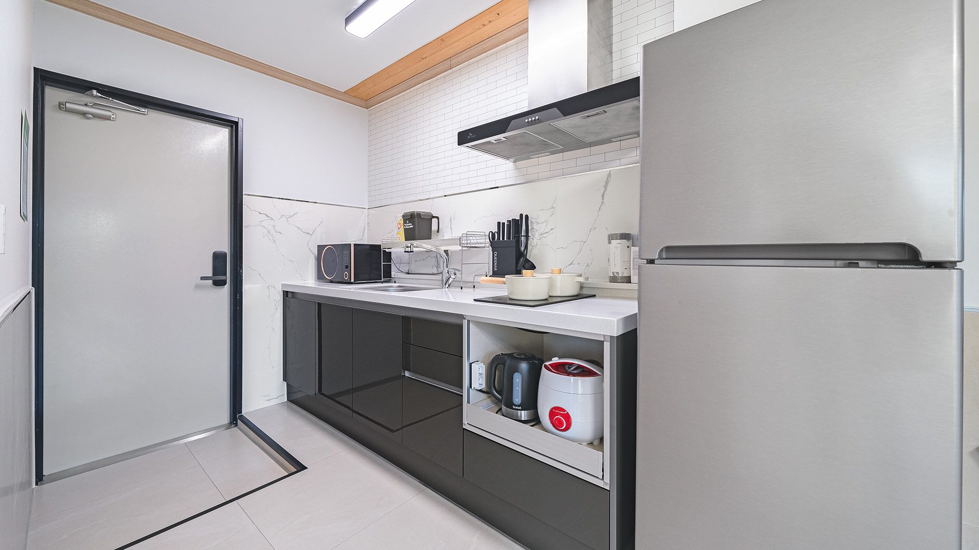 A modern kitchen with a built-in microwave, kettle, and coffee maker, a stainless steel refrigerator, and a door with a handle.