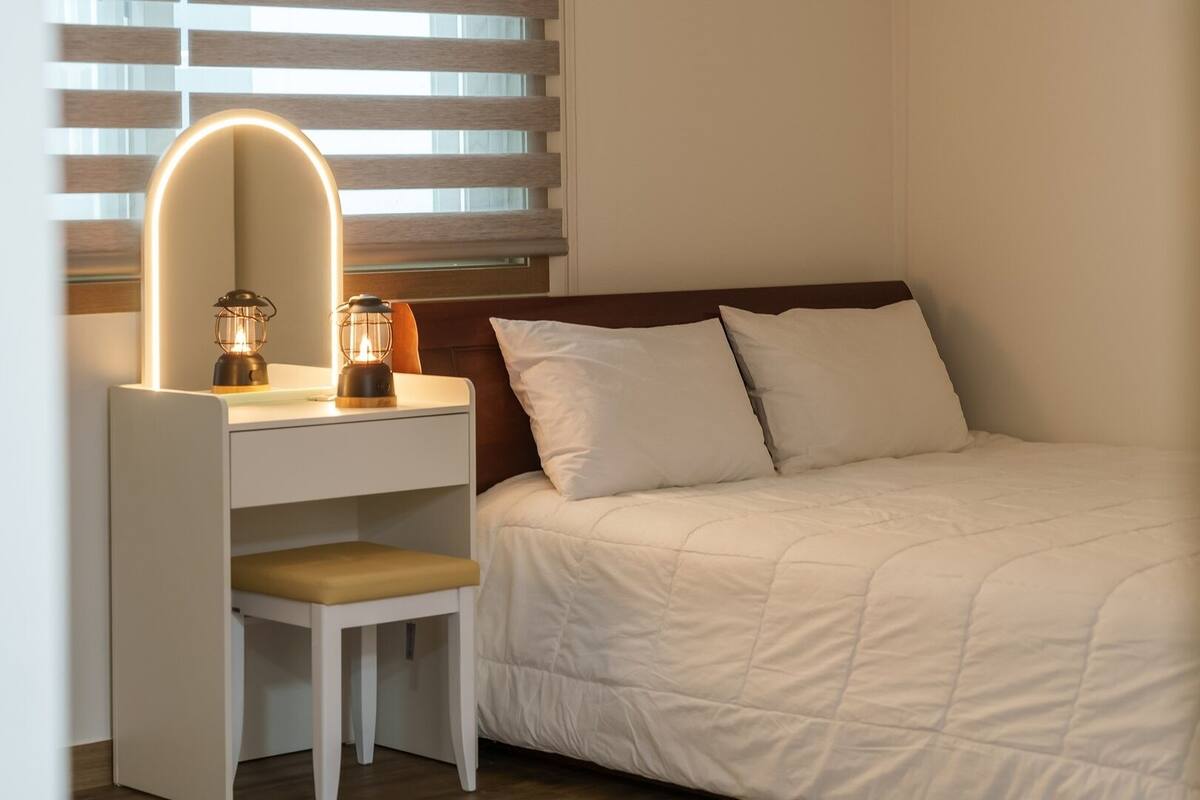 A bedroom with a bed, a desk with a chair, a mirror, and two wall-mounted lamps.