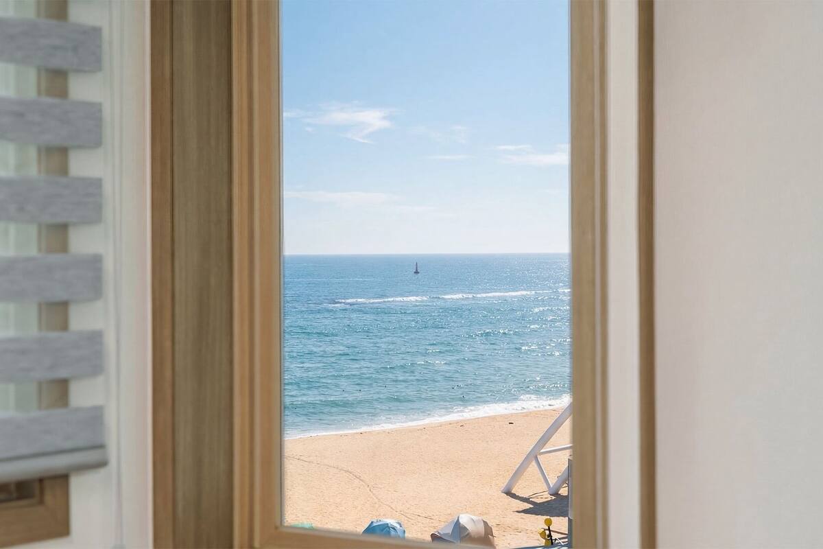 A window view of a beach with a chair and a hat on the sand.