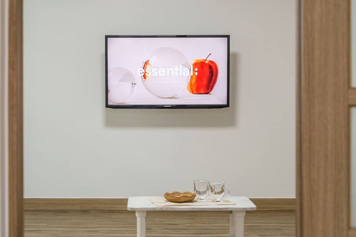 A flat-screen TV mounted on a wall, a small white table with a basket and two glasses, and a wooden floor.