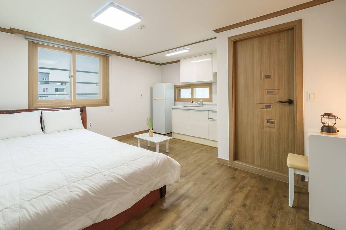 A bedroom with a bed, a small table, a window, a door, and a kitchenette in the background.