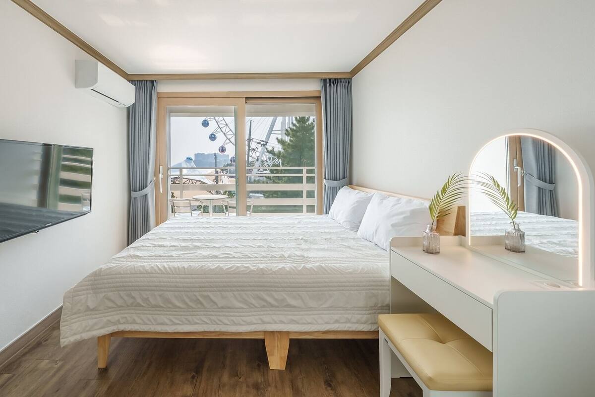 A bedroom with a bed, a TV mounted on the wall, a bedside table with a lamp, and a view of the outside.