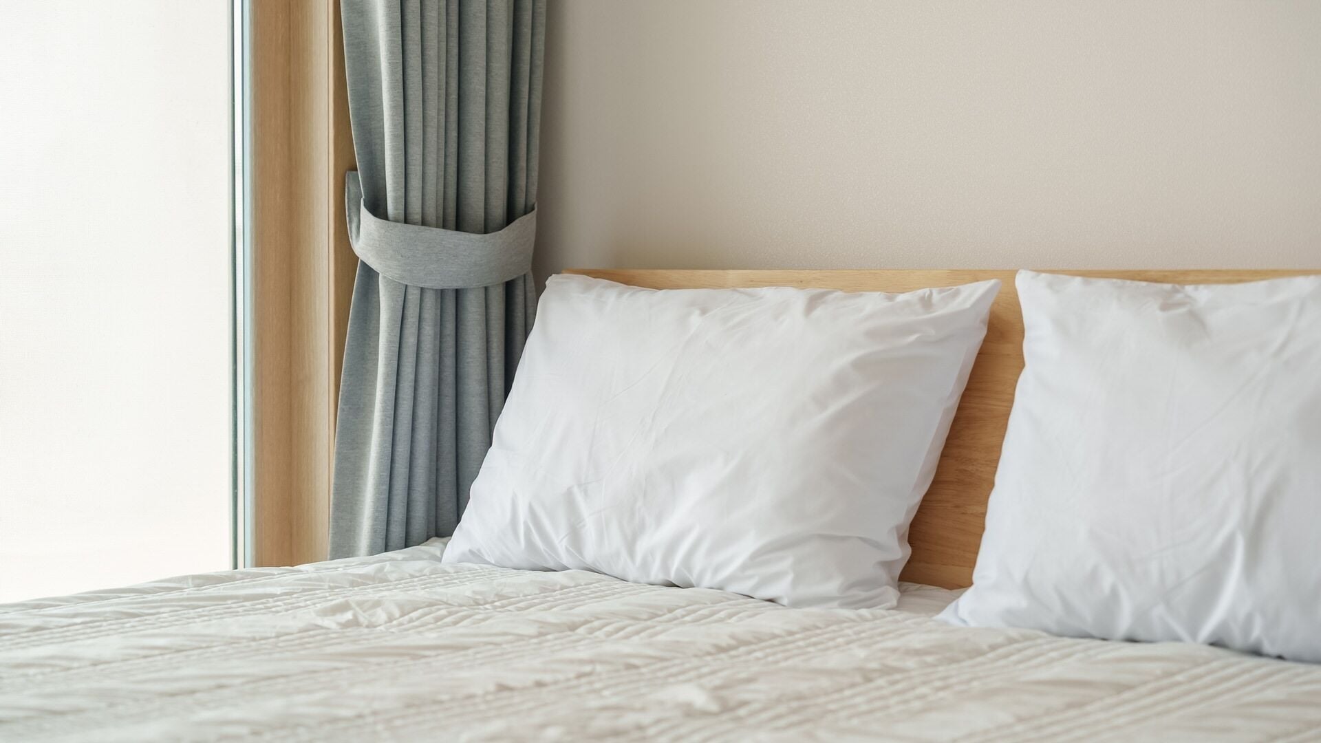 Een bed met witte beddengoed en kussens, een houten hoofdbord en een raam met grijze gordijnen.