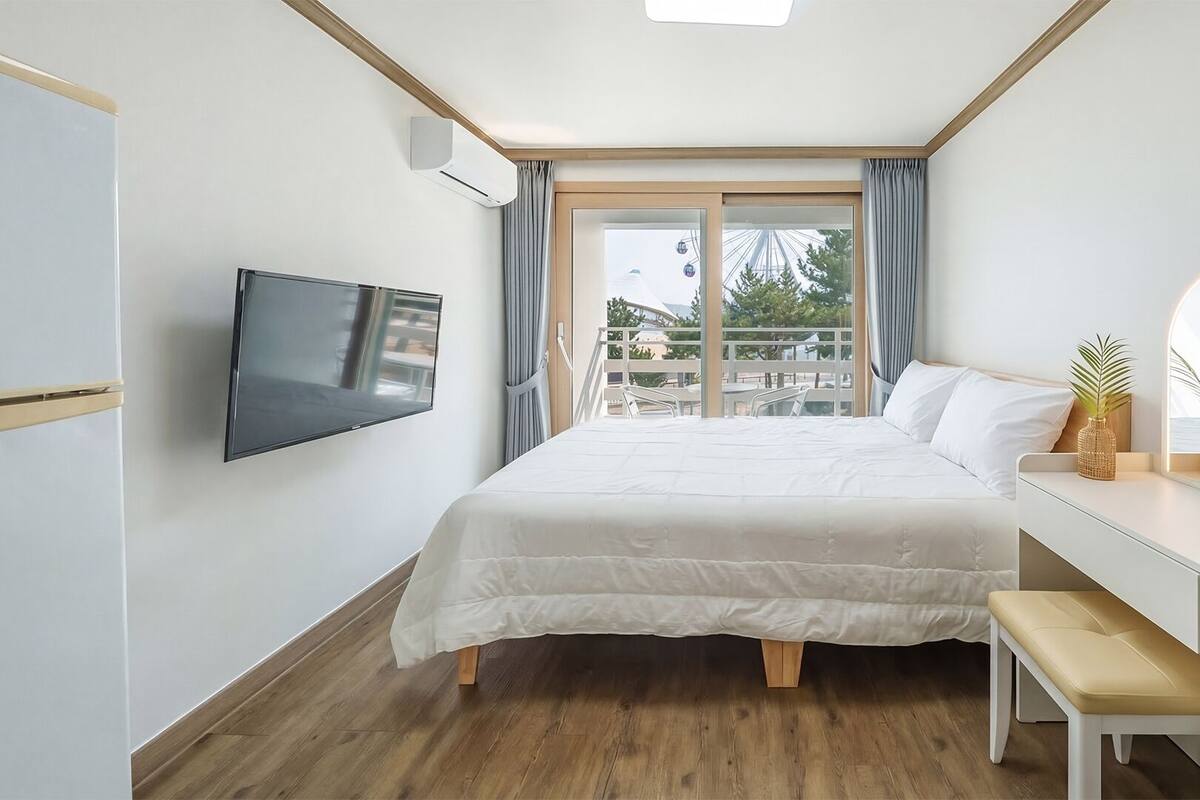 A hotel room with a bed, a TV, a desk with a mirror, and a balcony with a view of a Ferris wheel.
