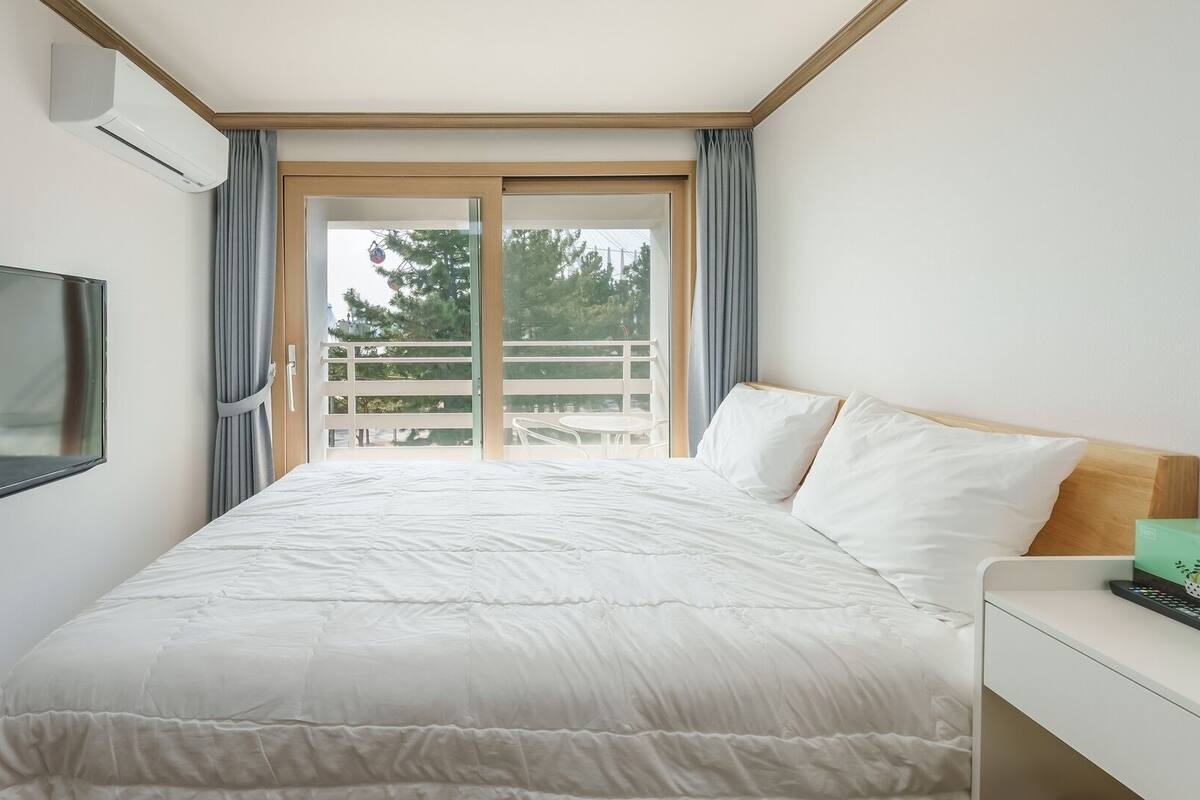 A hotel room with a bed, a TV, a desk, and a balcony with a view of trees.