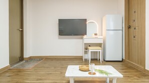 A minimalist room with a flat-screen TV, a white desk with a chair, a small table with a glass and a woven basket, and a white refrigerator.