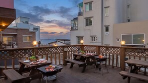 Eine Dachterrasse mit Holztischen und einem Grill, mit Blick auf eine Stadtlandschaft im Abendrot.