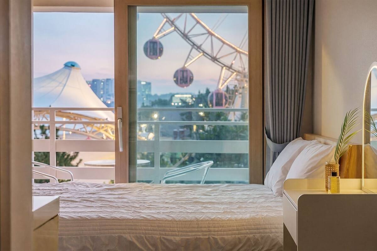 A hotel room with a bed, a bedside table with a mirror, and a view of a Ferris wheel.