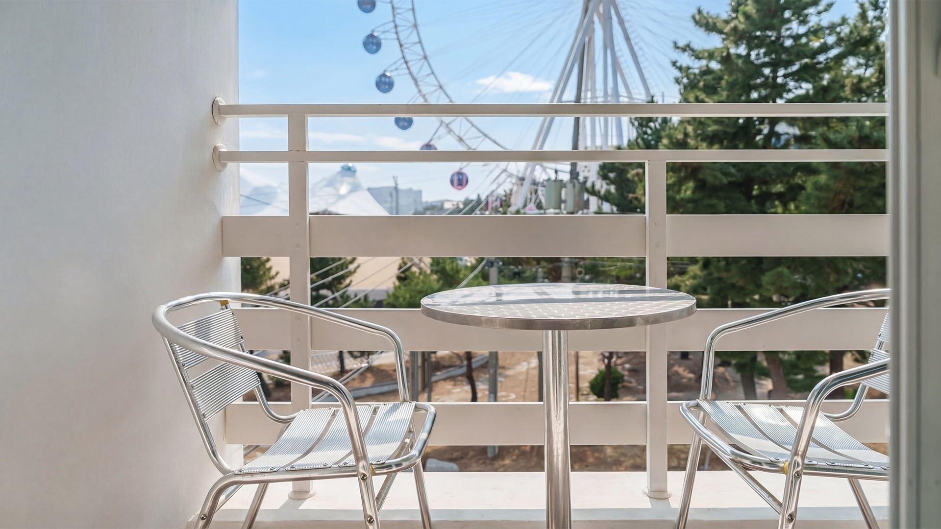 A balcony with a round table and two metal chairs, overlooking a Ferris wheel and trees.